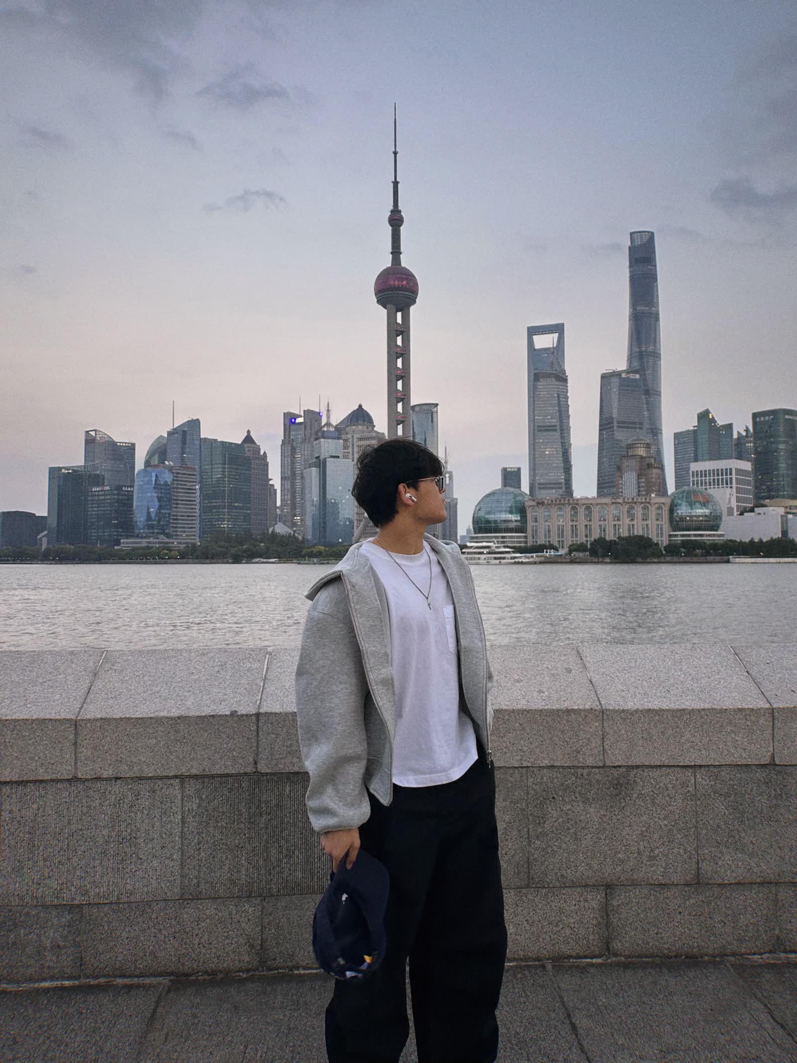 A person on a riverside promenade looking across the water at a city skyline at dusk.