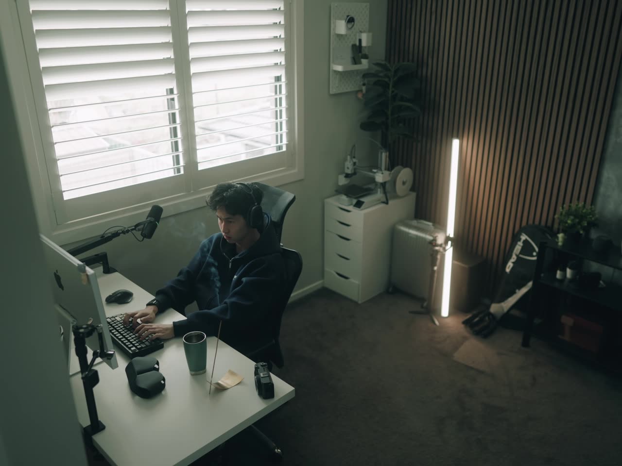 A person in headphones at a desk beside a shuttered window, an LED tube lit behind.