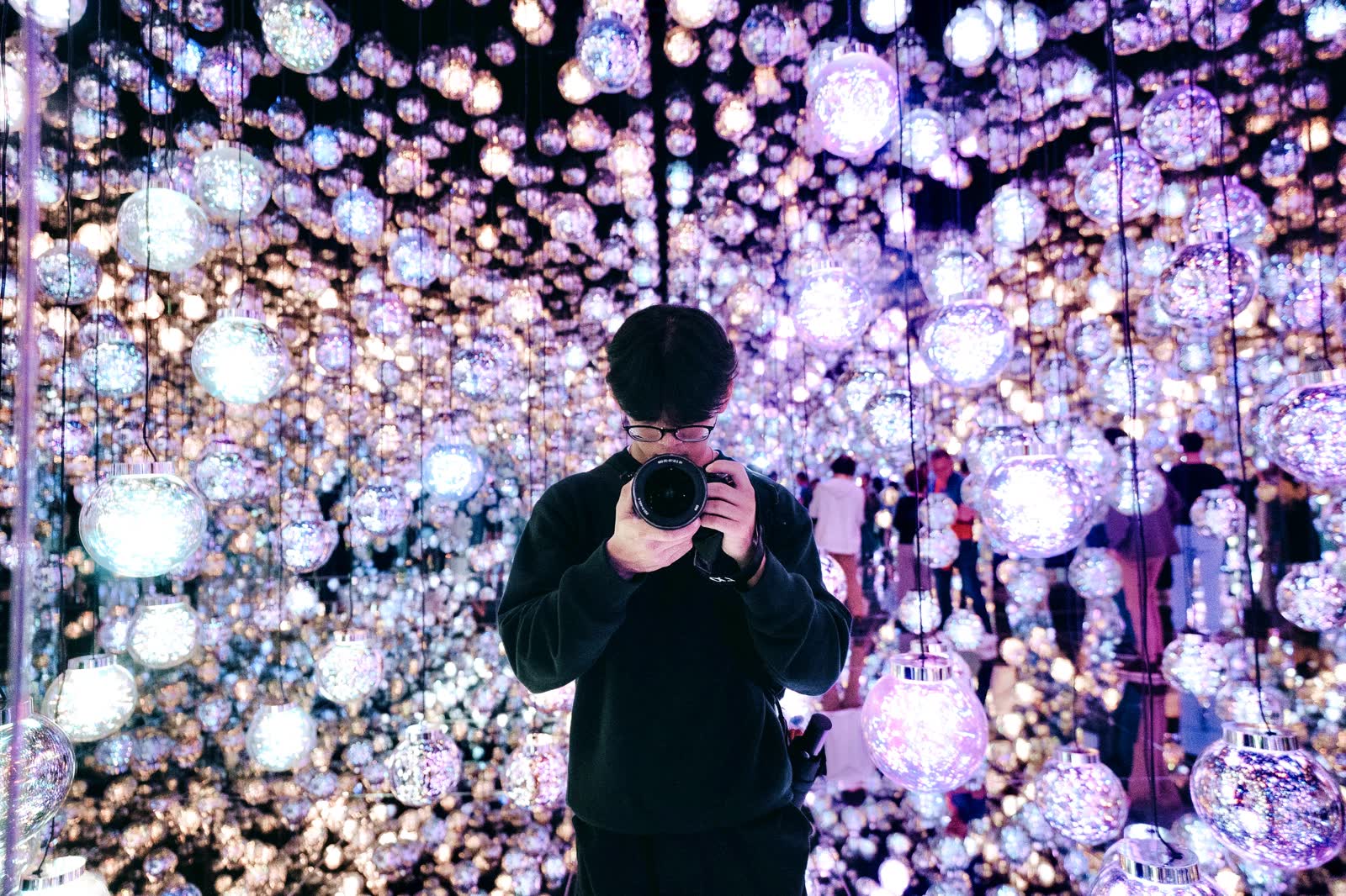 A photographer holding a camera to his eye inside a room of hanging mirrored lights.